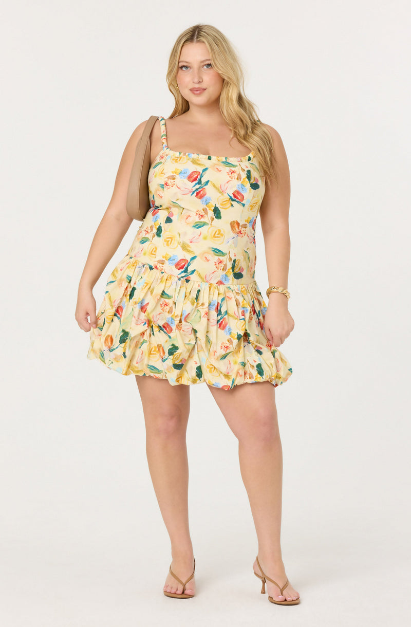 A woman is wearing a sleeveless, knee-length dress with a colorful, floral pattern on a yellow background. The dress has a fitted bodice with thin straps and a ruffled, gathered skirt. She is also holding a tan handbag over one shoulder, wearing a bracelet, and clear high-heeled sandals.