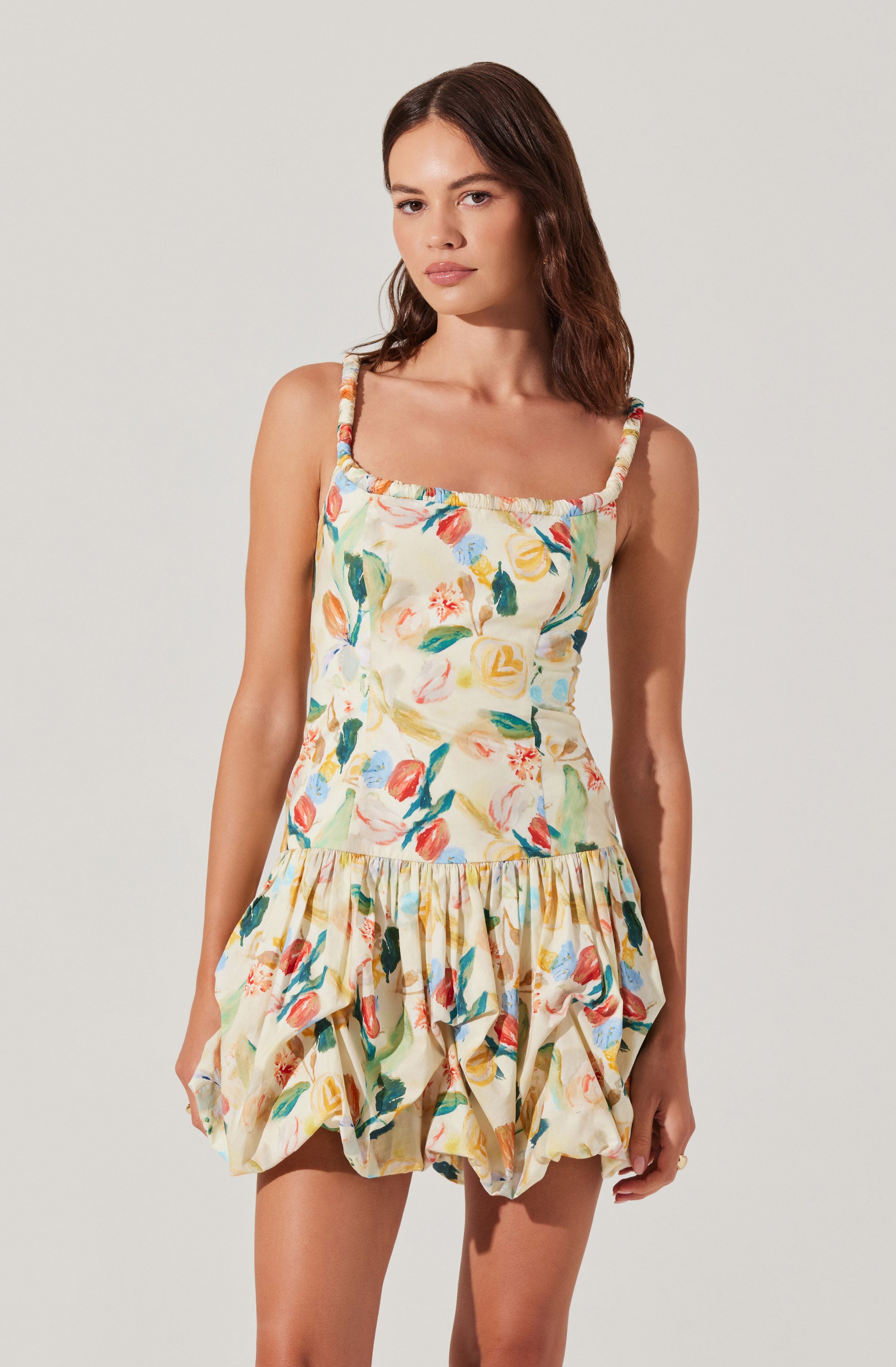 A young woman is wearing a sleeveless, knee-length dress with wide, ruffled straps. The dress features a colorful, abstract floral print on a light background, with a fitted bodice and a gathered, voluminous skirt that has a bubble hem.