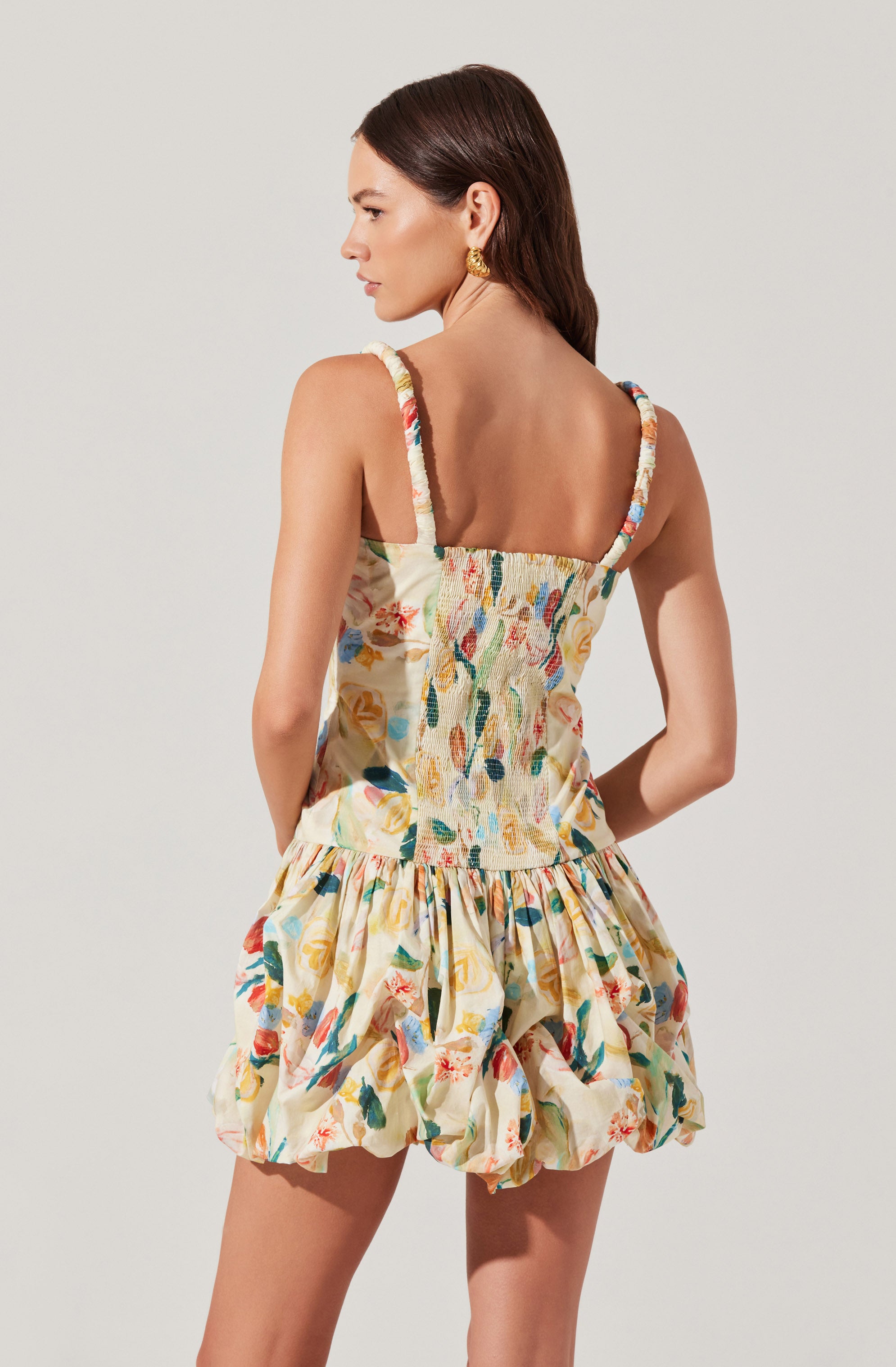 A woman is wearing a sleeveless romper with colorful, abstract floral patterns. The top has shirred detailing and wide shoulder straps, while the skirt portion features a ruffled, voluminous design. The fabric appears lightweight and suitable for warm weather.