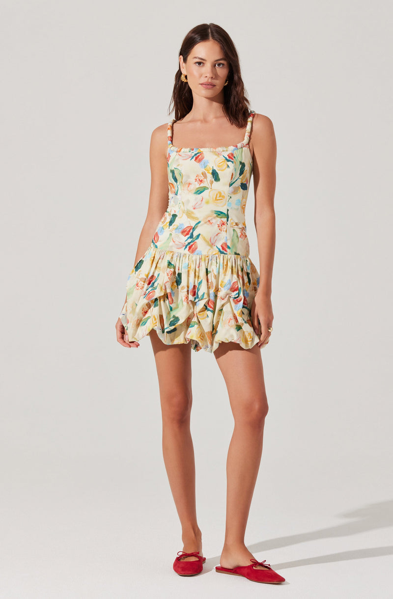 A woman is wearing a sleeveless, knee-length dress with a colorful floral print. The dress has a fitted bodice with spaghetti straps that feature ruffle details, and a ruffled, gathered skirt. She pairs the outfit with red flats that have bows on the front.