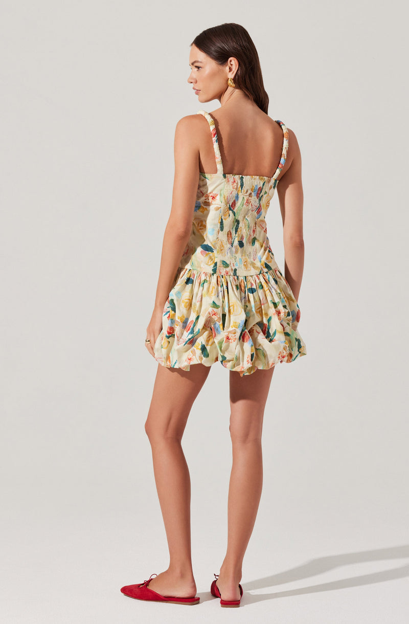 A woman is wearing a sleeveless, knee-length dress with wide straps. The dress has a fitted bodice with a smocked waist and a voluminous, ruffled skirt. The fabric features a colorful, abstract floral print in shades of yellow, orange, green, blue, and red. She is also wearing red flat shoes with a bow detail.