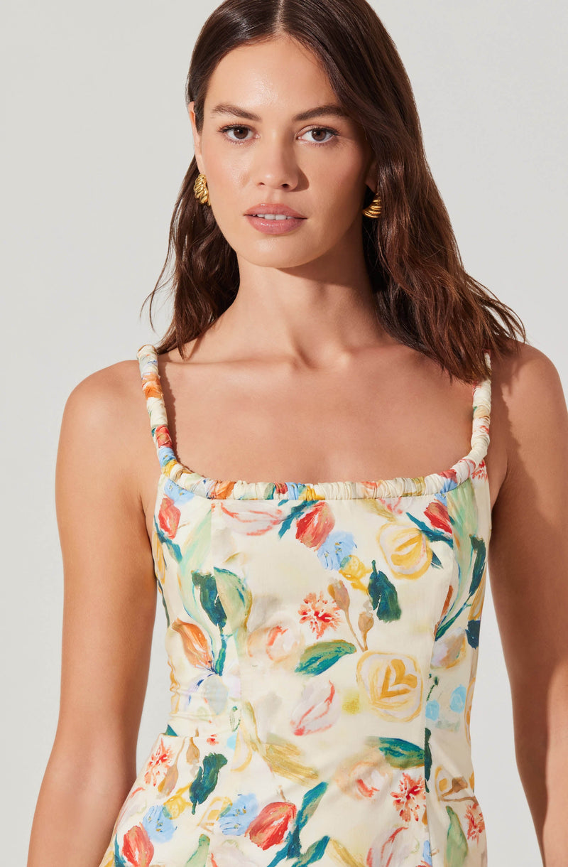 A woman is wearing a sleeveless dress with thin, gathered straps and a floral pattern. The dress features a variety of colorful flowers and leaves in shades of red, yellow, blue, green, and orange on a light, possibly cream or off-white background. She accessorizes with gold earrings.