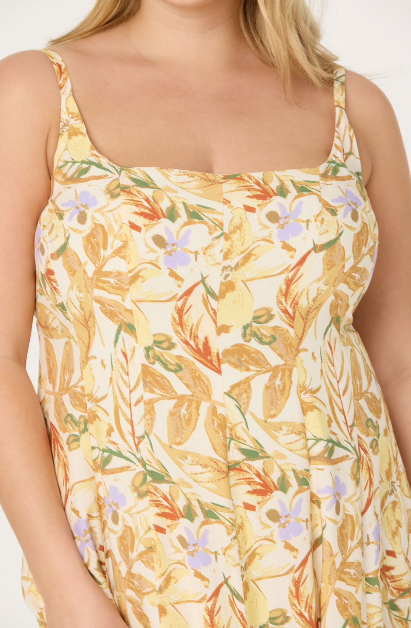 A woman is wearing a sleeveless dress with spaghetti straps featuring a tropical floral pattern. The dress has a square neckline and is decorated with yellow, green, orange, and light purple floral and leaf designs on a light background.
