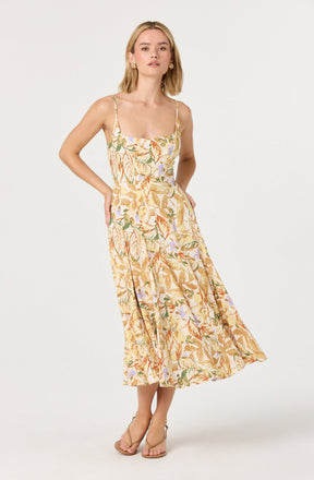 A woman is wearing a sleeveless, ankle-length dress with a fitted bodice and thin spaghetti straps. The dress features a light background with a colorful floral and leaf pattern in shades of orange, green, purple, and beige. She is also wearing beige open-toe high-heeled sandals.