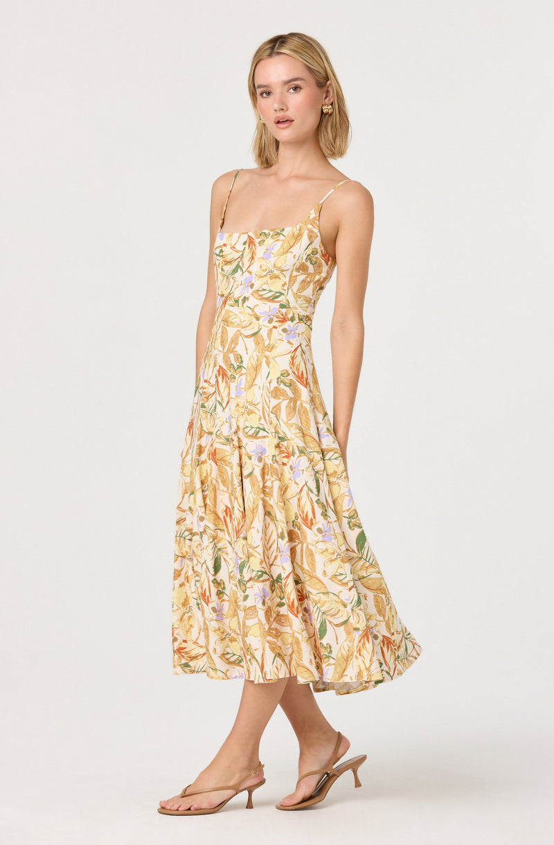 A woman is wearing a sleeveless, midi-length dress with a fitted bodice and thin spaghetti straps. The dress has a light, floral print pattern with shades of yellow, orange, green, and purple on a pale background. She is also wearing beige open-toe heels with a thin strap across the toes and a small heel.