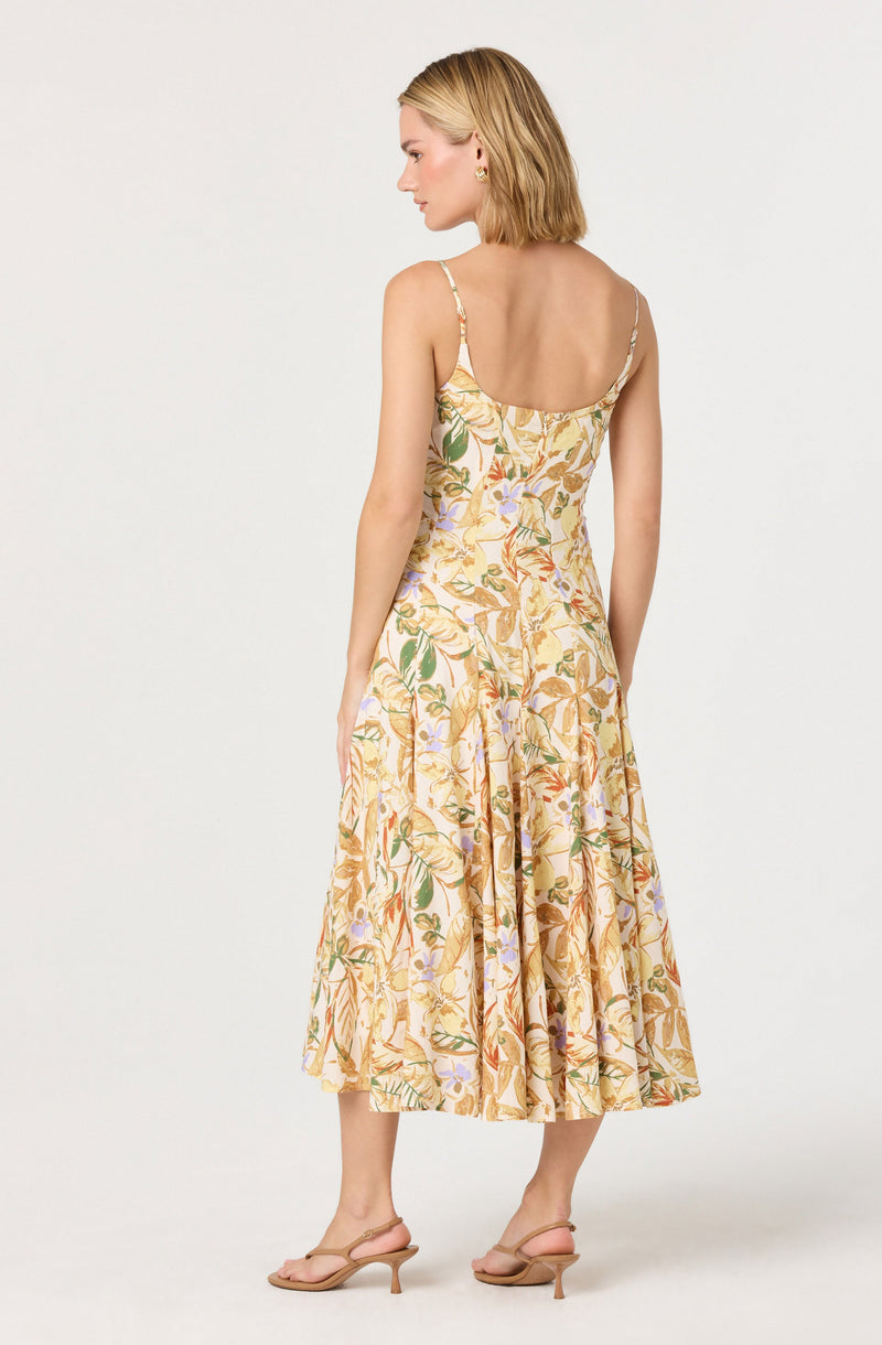 A woman is wearing a sleeveless, spaghetti strap dress with a floral pattern in shades of yellow, orange, green, and light purple. The dress has a fitted bodice and a flowing, mid-length skirt. She is also wearing beige, open-toe, low-heeled sandals.