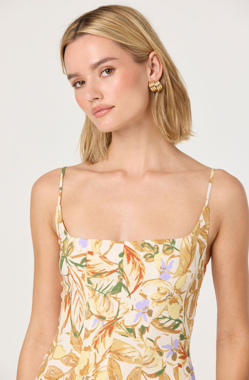 A woman is wearing a sleeveless dress with thin spaghetti straps. The dress features a colorful, floral pattern with shades of yellow, orange, green, and purple on a light background. She is also wearing large, gold, twisted hoop earrings.