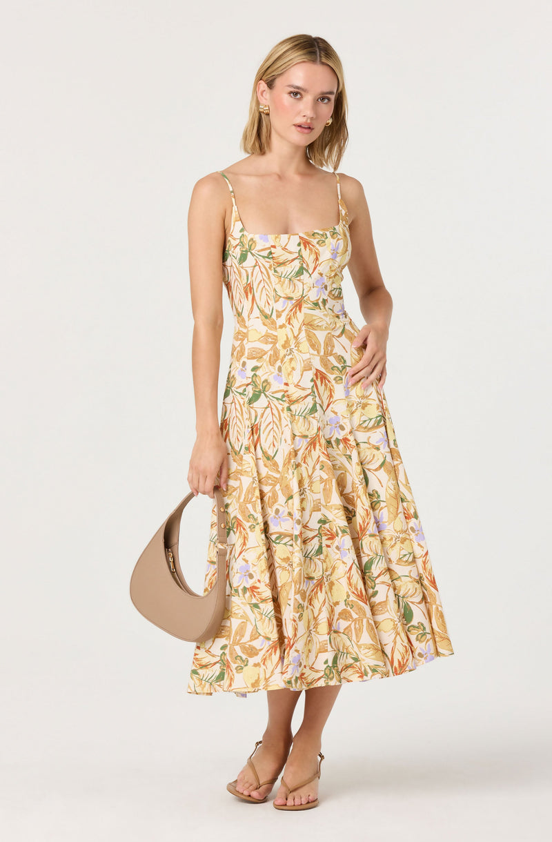 A woman is wearing a sleeveless, midi-length dress with a floral and leaf pattern in shades of yellow, orange, green, and purple on a light background. The dress has thin spaghetti straps and a fitted bodice that flares out at the skirt. She is holding a beige, curved handbag in her right hand and wearing open-toe, ankle-strap high heels.
