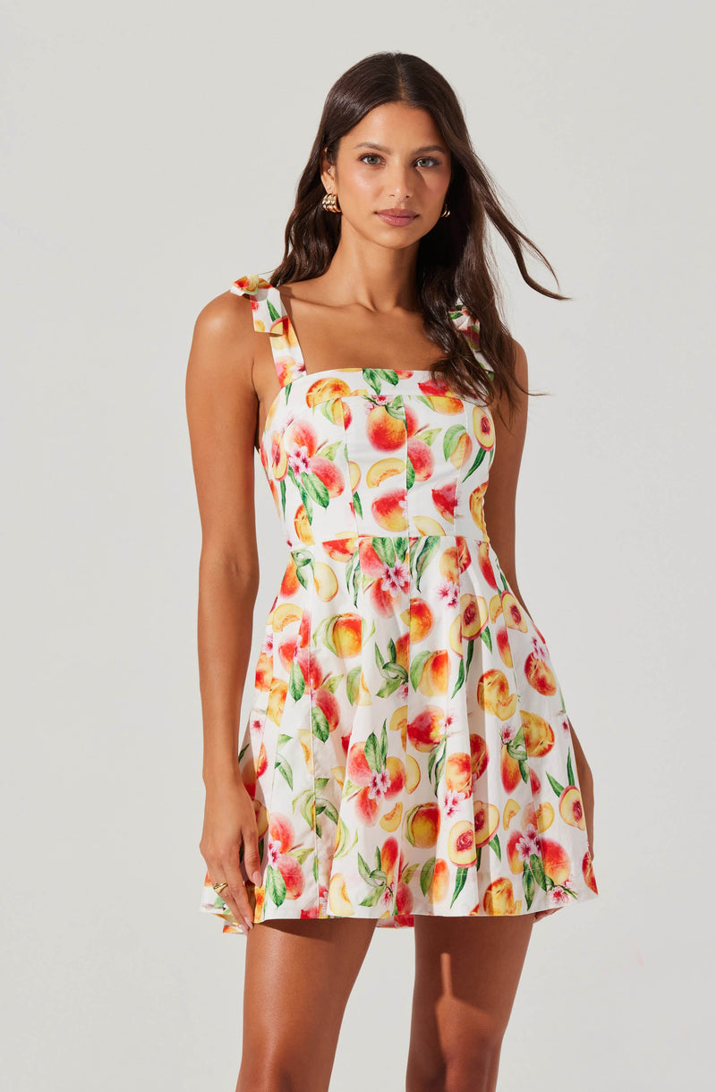 A woman is wearing a sleeveless, knee-length dress with a square neckline and shoulder straps tied into bows. The dress features a colorful print of peaches, pink flowers, and green leaves on a white background.