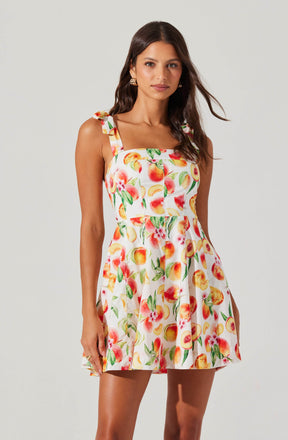 A woman is wearing a sleeveless, knee-length dress with a square neckline and shoulder straps tied into bows. The dress features a colorful print of peaches, pink flowers, and green leaves on a white background.