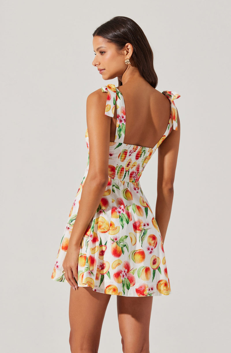 A woman is wearing a sleeveless summer dress with shoulder straps tied into bows. The dress features a vibrant fruit print, including peaches, apples, and pomegranates, on a light background. The fitted bodice has a shirred detail, and the skirt flares out slightly.