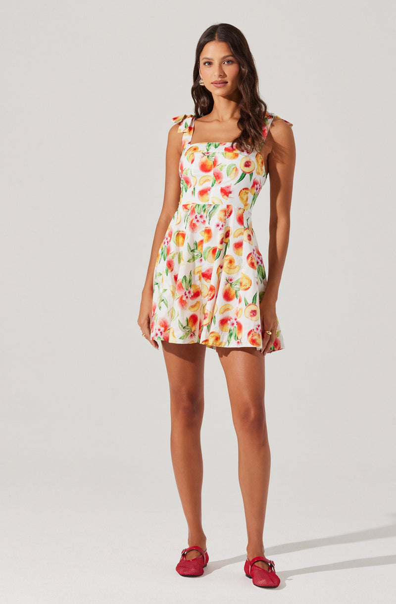 A woman is wearing a sleeveless, above-the-knee dress with a square neckline and shoulder tie straps. The dress features a colorful fruit print with peaches, apples, and lemons on a white background. She is also wearing pink shoes.