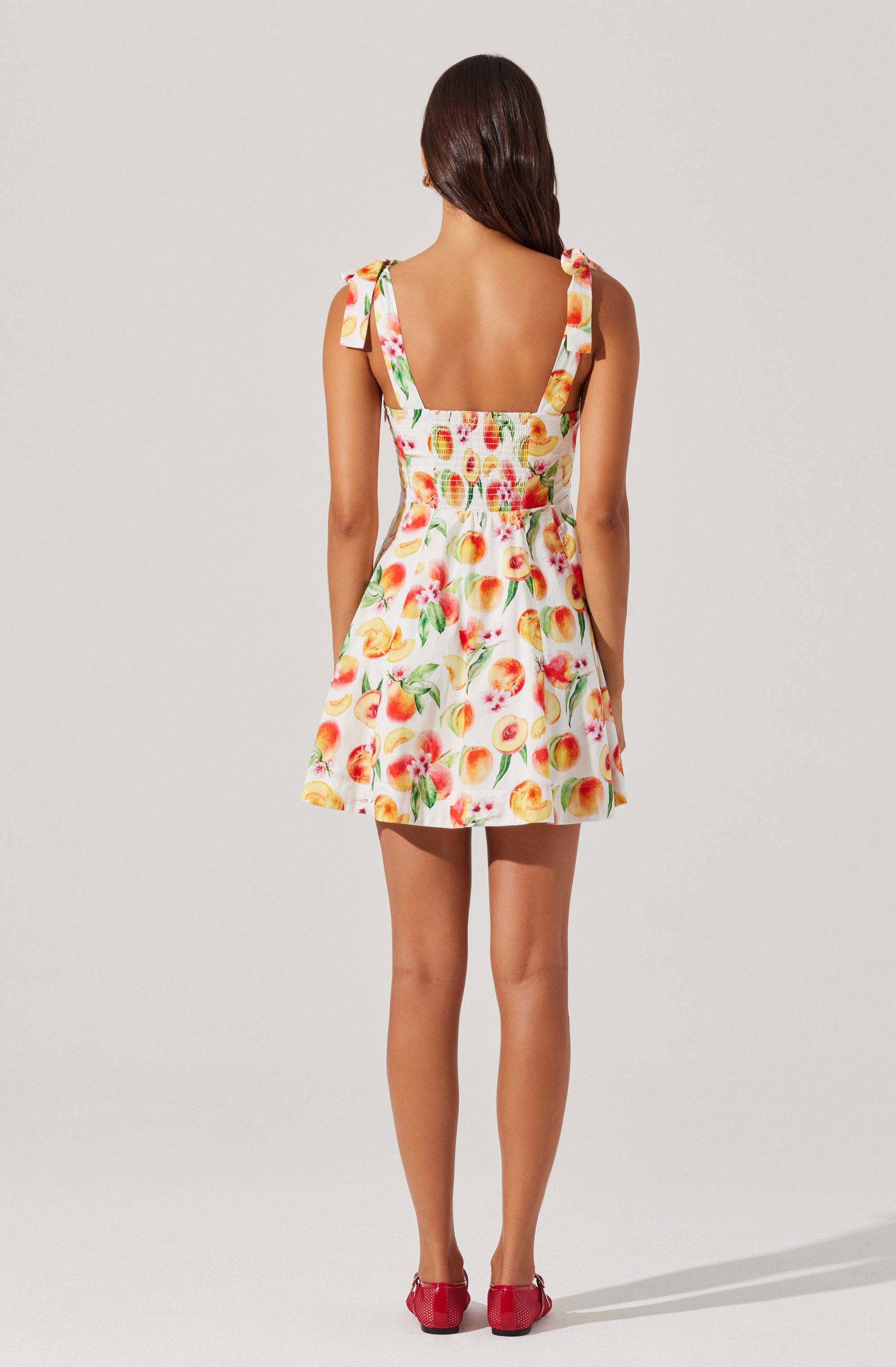 A woman is wearing a sleeveless, above-the-knee dress with a colorful fruit pattern, featuring peaches and other fruits. The dress has wide shoulder straps tied into bows. She is also wearing red shoes.