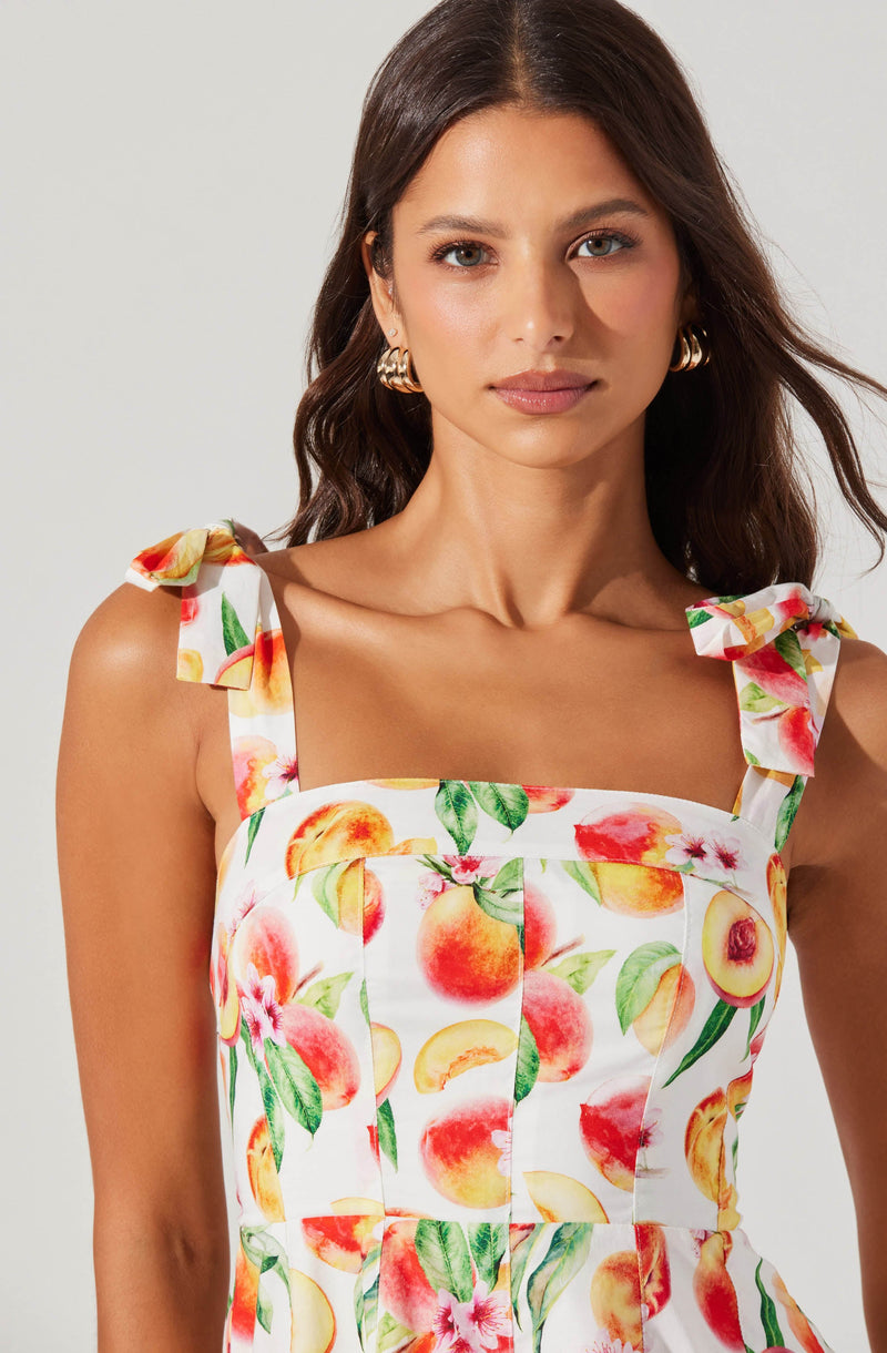 A woman is wearing a sleeveless dress with shoulder straps tied into bows. The dress features a colorful print of peaches and peach blossoms on a white background.