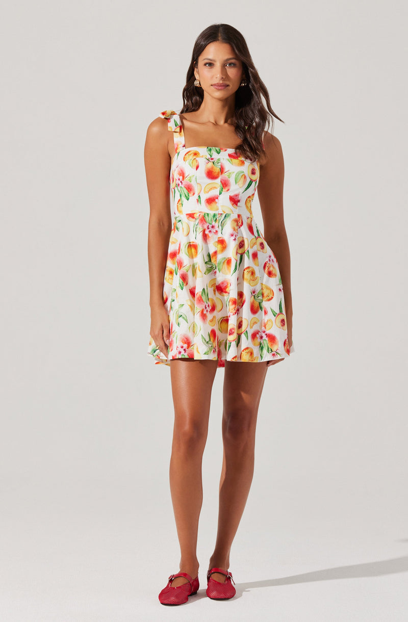A woman is wearing a sleeveless, above-the-knee dress with a square neckline and ruffled shoulder straps. The dress features a vibrant, colorful fruit print on a white background. She has paired the dress with red shoes.