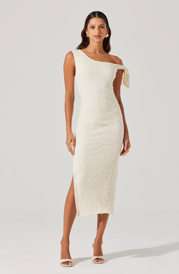 A woman is wearing a sleeveless, off-the-shoulder, ankle-length dress in a textured cream fabric. The dress features a unique strap detail on one shoulder that ties into a bow. She is also wearing open-toe heels and gold earrings.