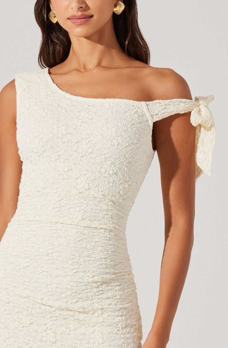 A woman is wearing a sleeveless, cream-colored textured dress with an asymmetrical neckline. The dress features a decorative bow on the shoulder strap.