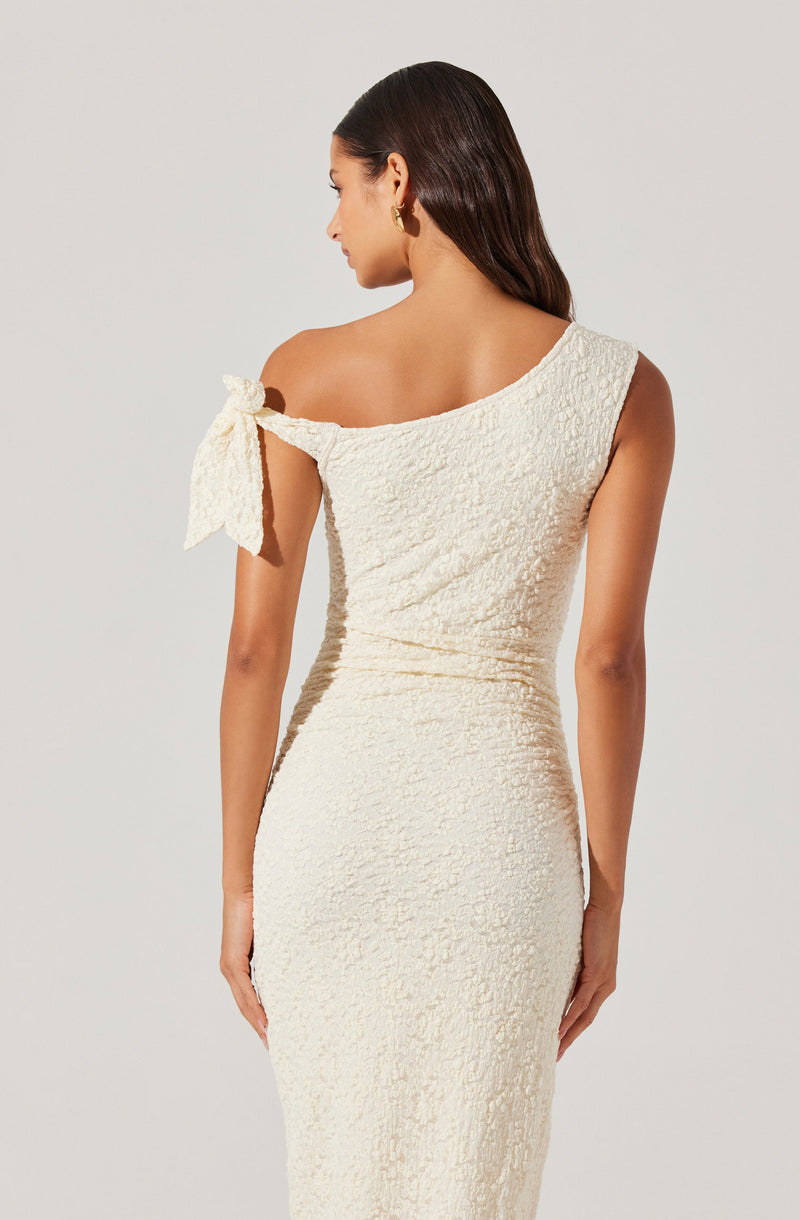 A woman is wearing a one-shoulder, cream-colored dress with a textured, lace-like fabric. The dress has a decorative bow on the shoulder. She is accessorized with gold hoop earrings.