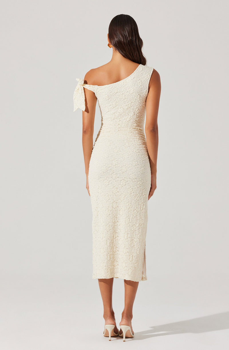 A woman is wearing a midi-length, off-the-shoulder dress in a textured, cream-colored fabric. One shoulder features a strap with a bow detail, while the other shoulder is strapless. The dress is fitted and extends to below the knees, with a slight slit on the side. She is also wearing high-heeled shoes.