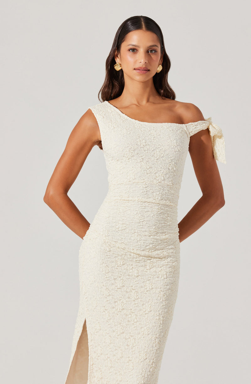A woman is wearing a white, textured, fitted dress with an asymmetrical neckline. The dress features a single shoulder with a decorative bow on the shoulder strap and a slit on the side. She accessorizes with large, round gold earrings.