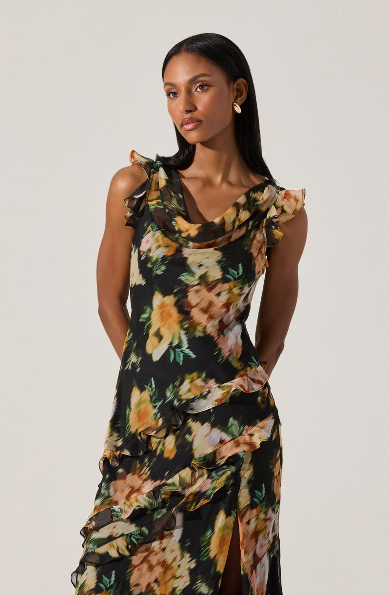 A woman is wearing a sleeveless, floral dress with ruffled shoulder details and a draped neckline. The dress features a black background with large, colorful flower prints in shades of yellow, peach, pink, and green. The fabric has a flowing, layered design with ruffles along the bodice and skirt, and a slit in the front.