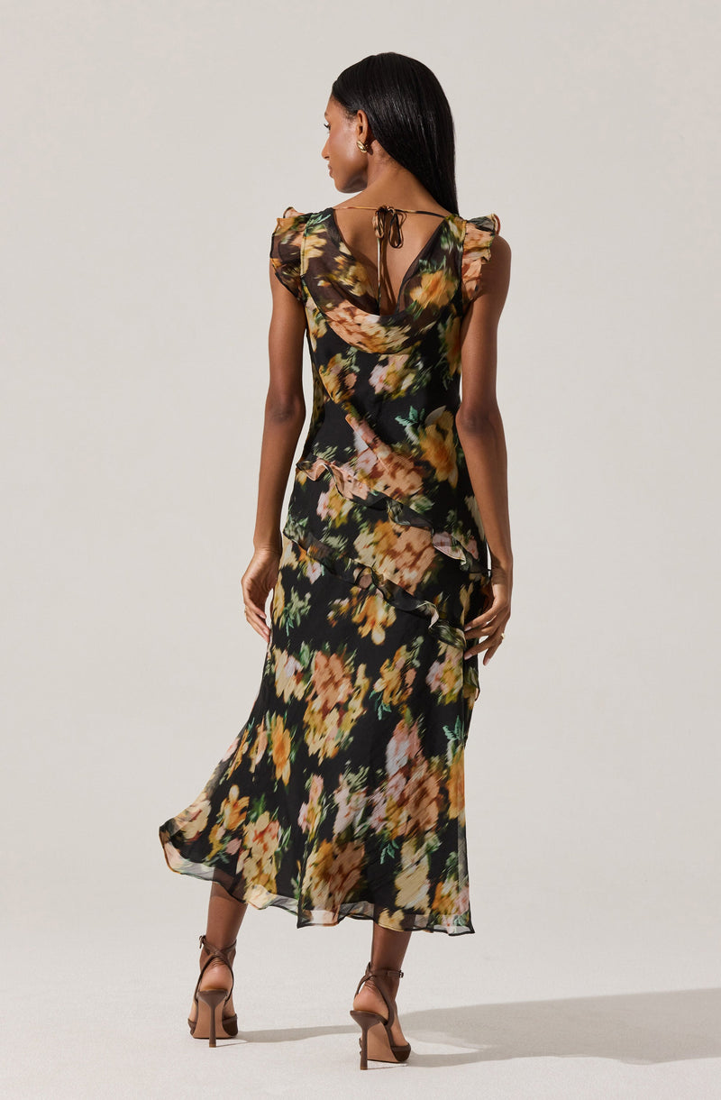 A woman is wearing a long, flowing dress with a black background and an abstract floral print in shades of yellow, orange, pink, and green. The dress features ruffled details along the sides and has shoulder pads. It has a tie at the back of the neck, creating a keyhole opening at the upper back. The fabric appears lightweight and semi-sheer. She is also wearing high-heeled sandals in a nude tone.