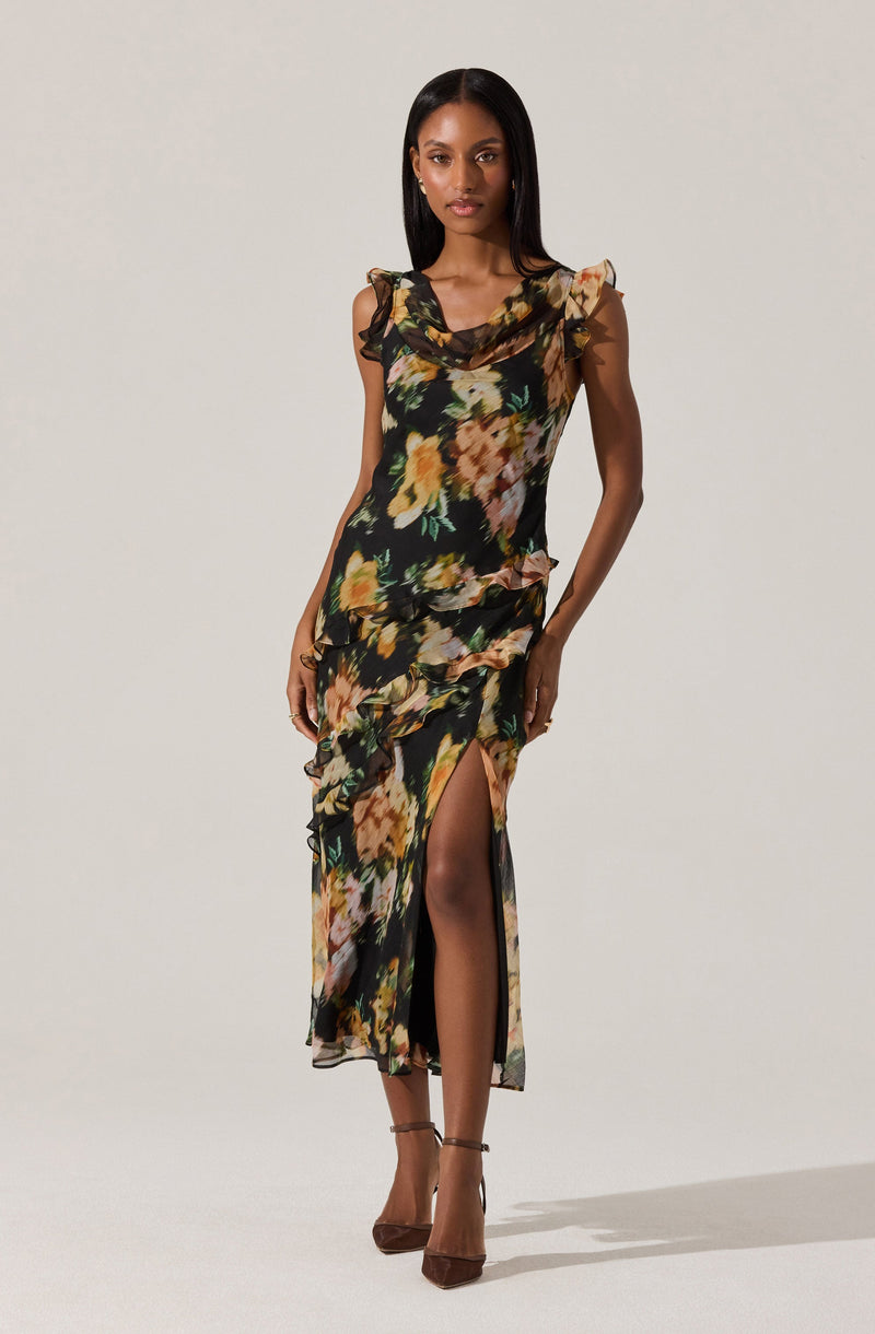 The dress is a sleeveless, floor-length gown with a black background and a floral pattern featuring shades of yellow, orange, green, and beige. It has ruffled accents along the shoulders and down the front, and a high slit on one side revealing the leg.