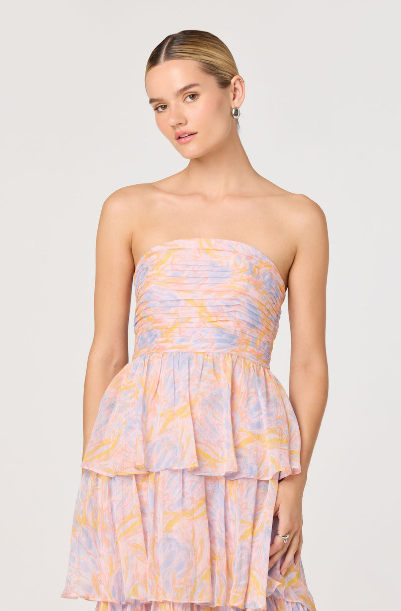 A woman is wearing a strapless, pastel-colored dress with a smocked bodice and a layered, ruffle skirt. The dress features a soft, abstract pattern in shades of pink, lavender, and yellow.