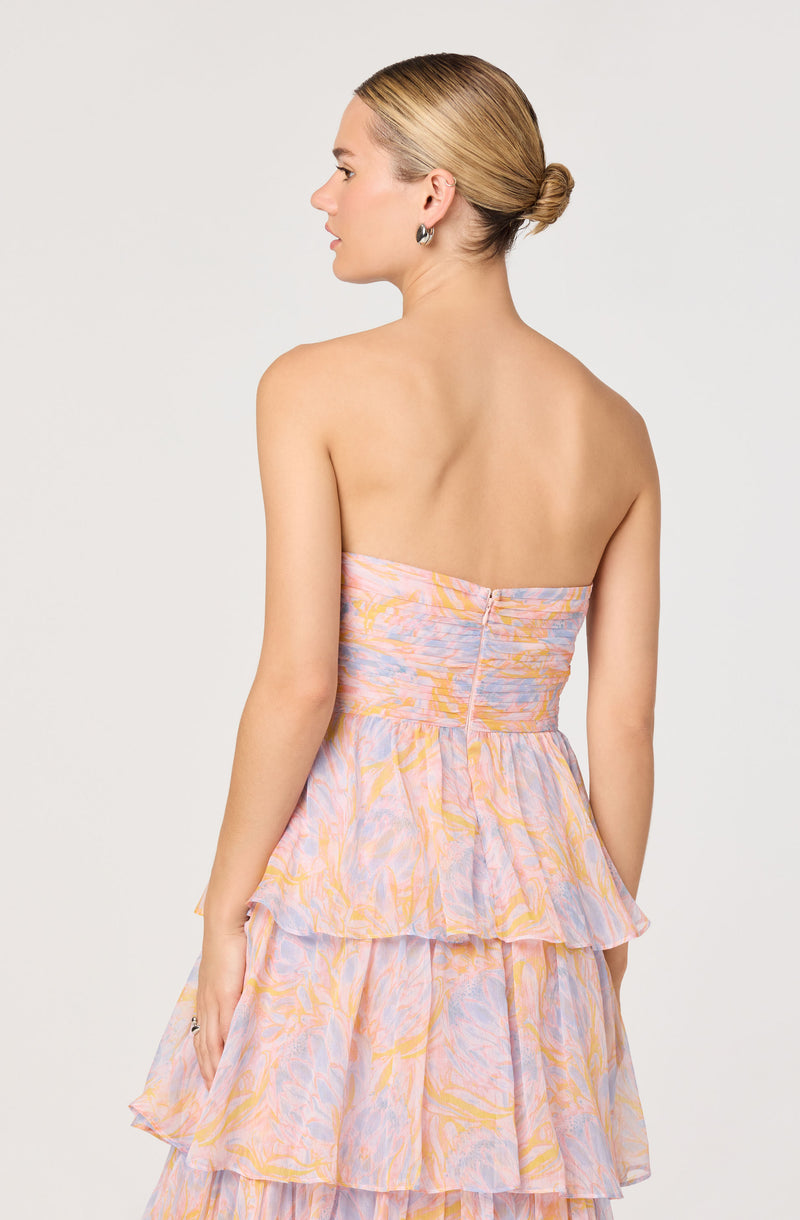 A woman is wearing a strapless dress with a pastel, abstract watercolor pattern in shades of pink, purple, yellow, and orange. The dress features a fitted bodice with a zipper closure at the back and a tiered, ruffled skirt with multiple layers.