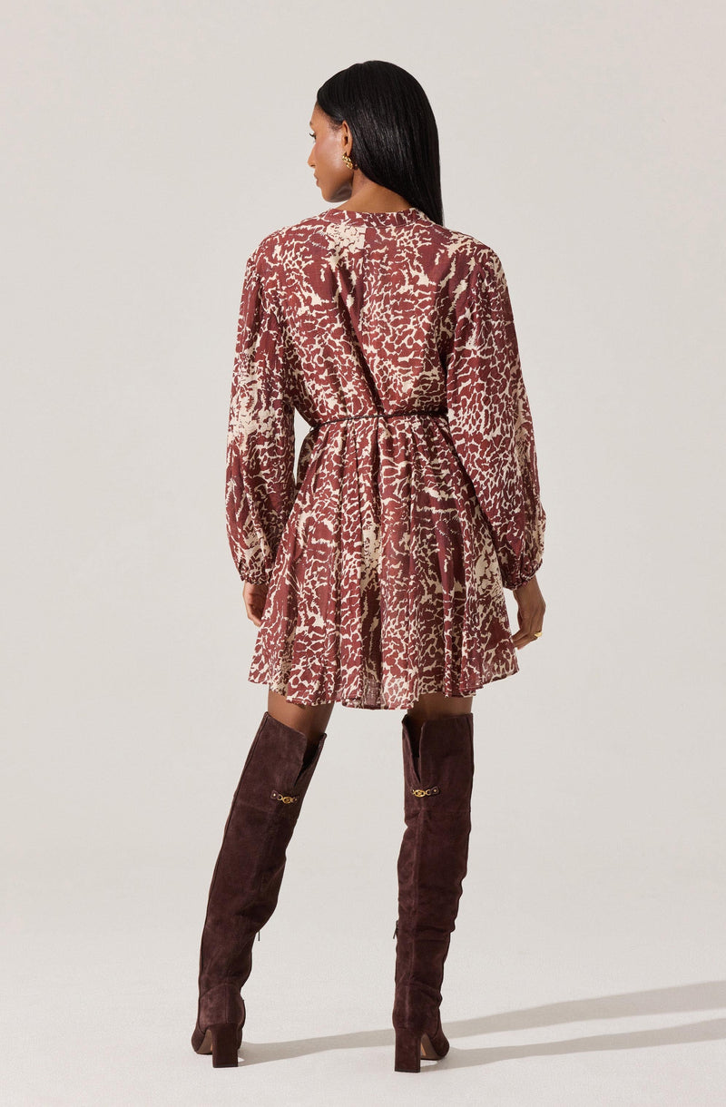The model is wearing a long-sleeved dress with a high neckline. The dress features an abstract, animal print-like pattern in shades of brown and beige. It has a cinched waist with a matching fabric belt and a flared, ruffled hem that falls above the knees. The dress appears to be made of a lightweight, flowy fabric. She is also wearing tall, dark brown suede over-the-knee boots with block heels.