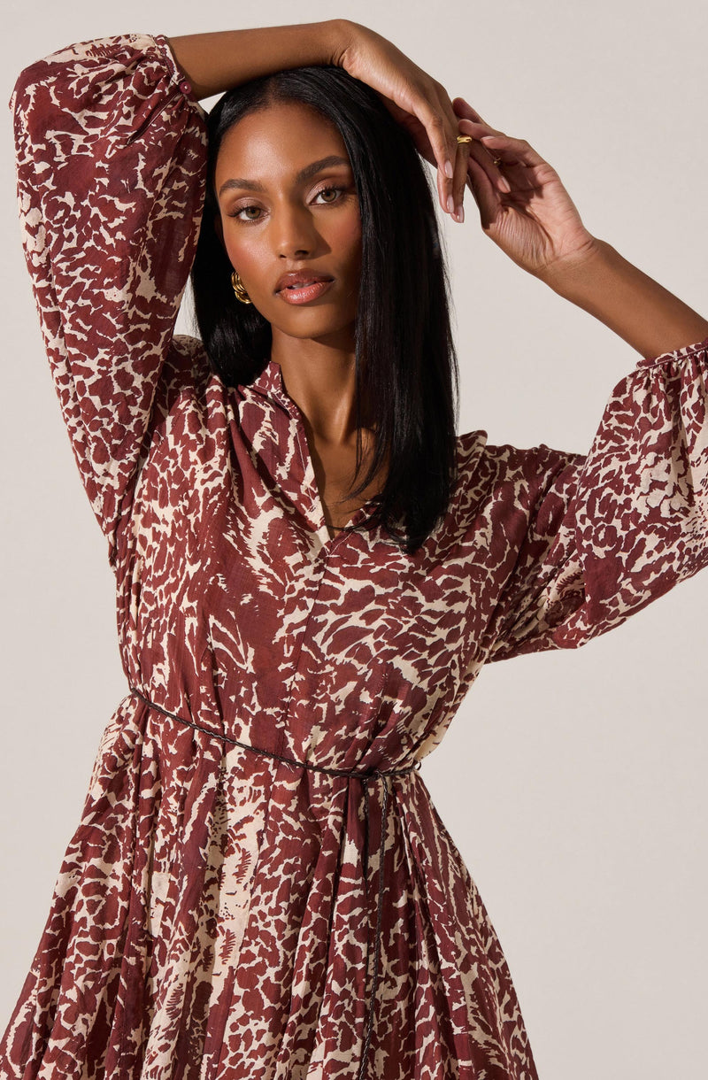 The dress features a loose, flowing fit with a collared neckline and long, slightly puffed sleeves that gather at the cuffs. It is printed with a bold, abstract animal print pattern in shades of reddish-brown and beige. The dress has a waist tie that cinches at the waist, creating a feminine silhouette.