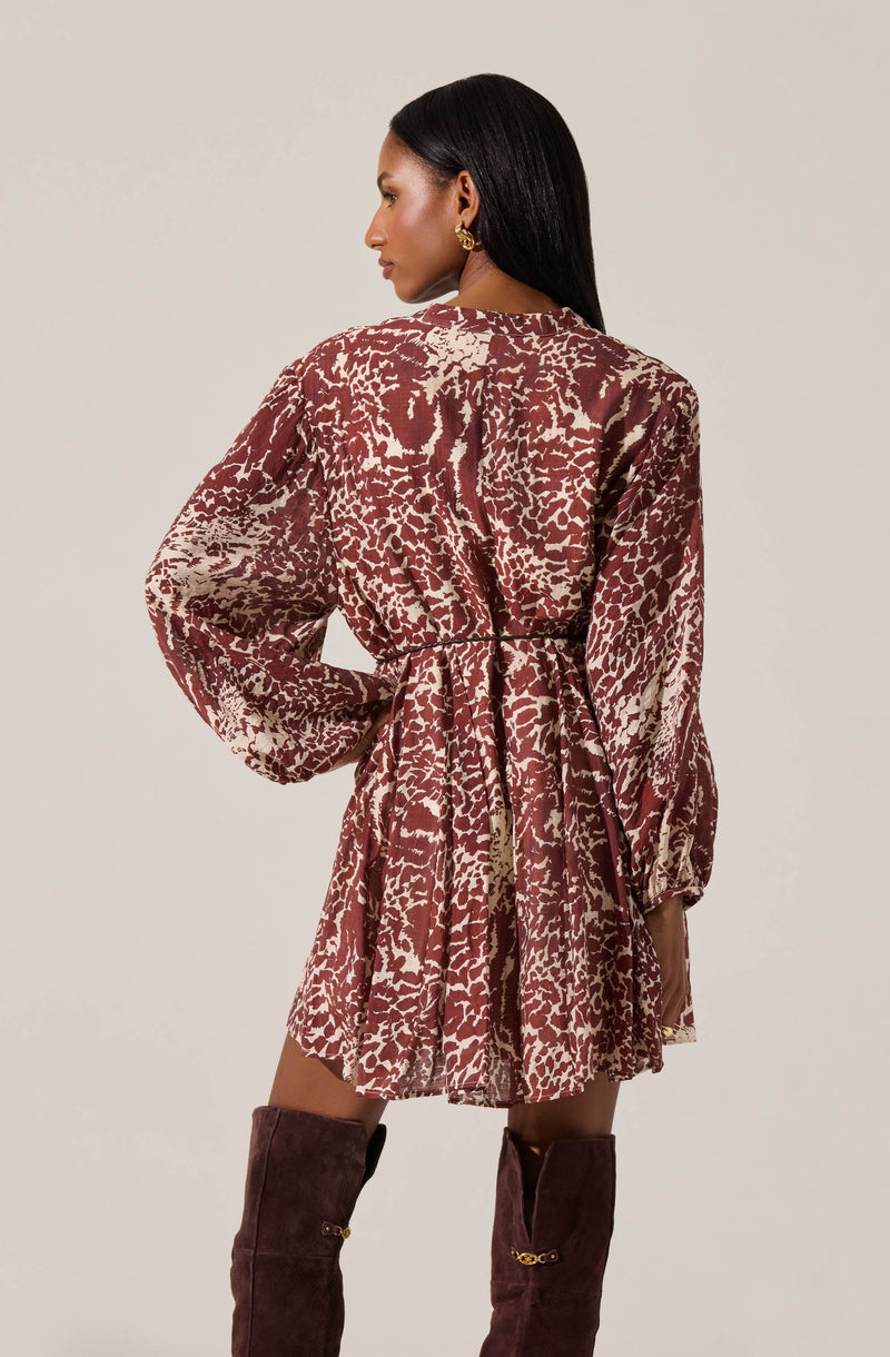 A woman is wearing a long-sleeved dress with a loose fit and a gathered waist. The dress has a bold, abstract animal print pattern in shades of beige and deep red. The sleeves are full and slightly puffed, with elastic cuffs.