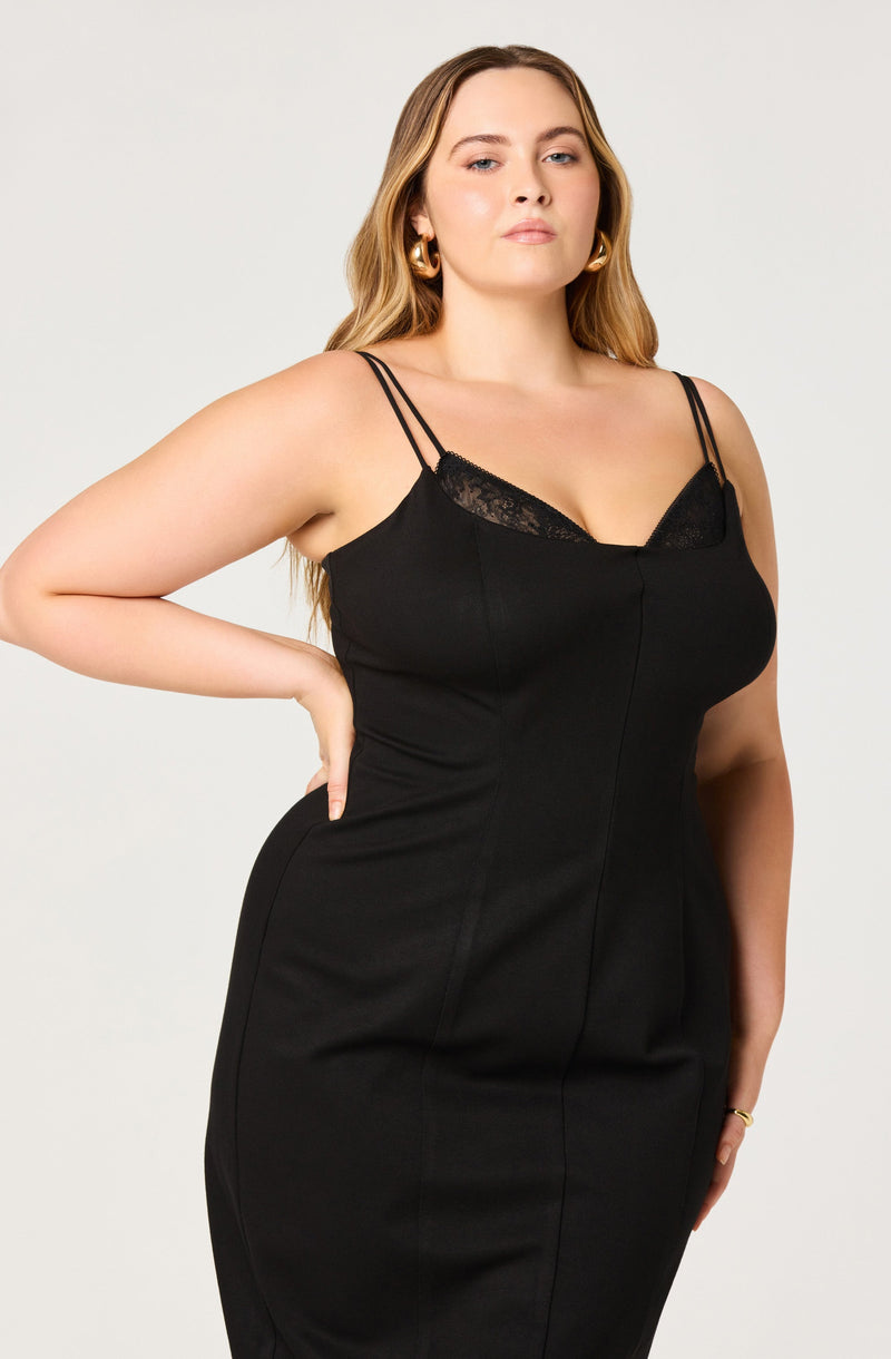 A woman is wearing a black sleeveless dress with spaghetti straps and lace detail at the neckline. The dress is fitted and has visible seam lines running down the front. She is accessorized with gold hoop earrings and a gold bracelet.