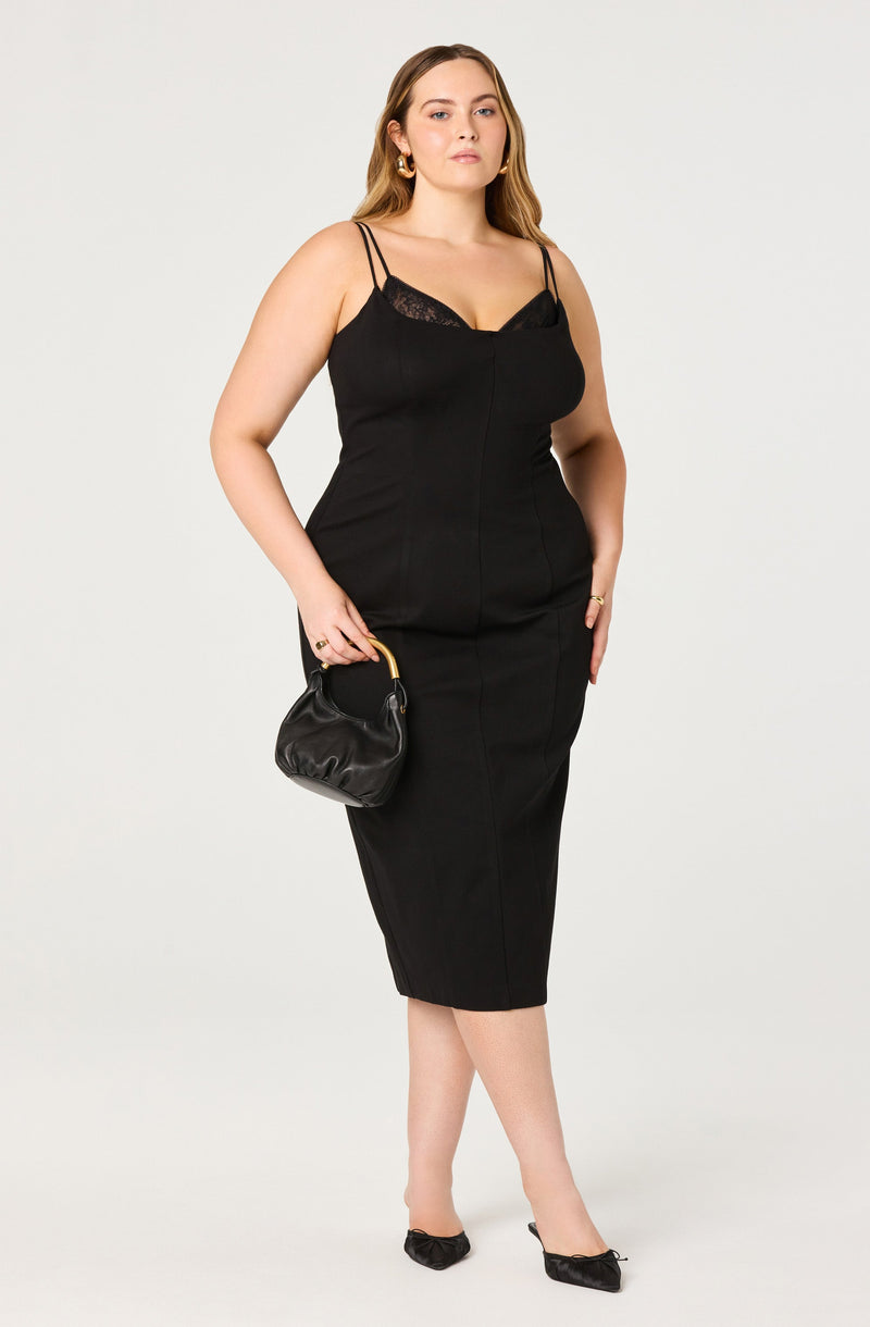 A woman wearing a fitted black midi dress with thin double straps and lace trim at the neckline. She is holding a small black handbag with a gold handle and is wearing black heels with bows.