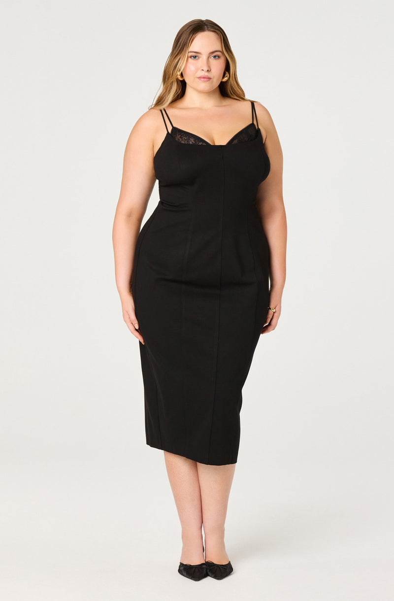 A woman is wearing a black sleeveless dress with double spaghetti straps and lace detail at the bust. The dress is fitted, midi-length, and has a sleek, elegant style. She is also wearing black shoes with a small bow detail.