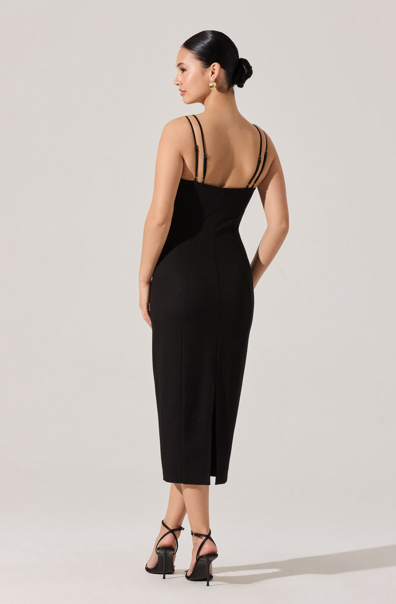 A woman is wearing a fitted, sleeveless black dress that extends below the knees and features a slit at the back. The dress has thin double straps across the shoulders, and she is accessorizing with gold earrings and black high-heeled sandals with ankle straps. Her hair is styled in a sleek bun.