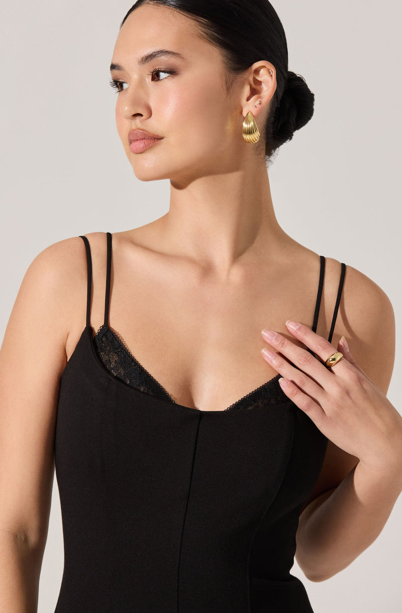 A woman is wearing a black dress with thin spaghetti straps and a lace trim at the neckline. She has gold hoop earrings and a gold ring on her left hand. Her hair is styled in a sleek, low bun.