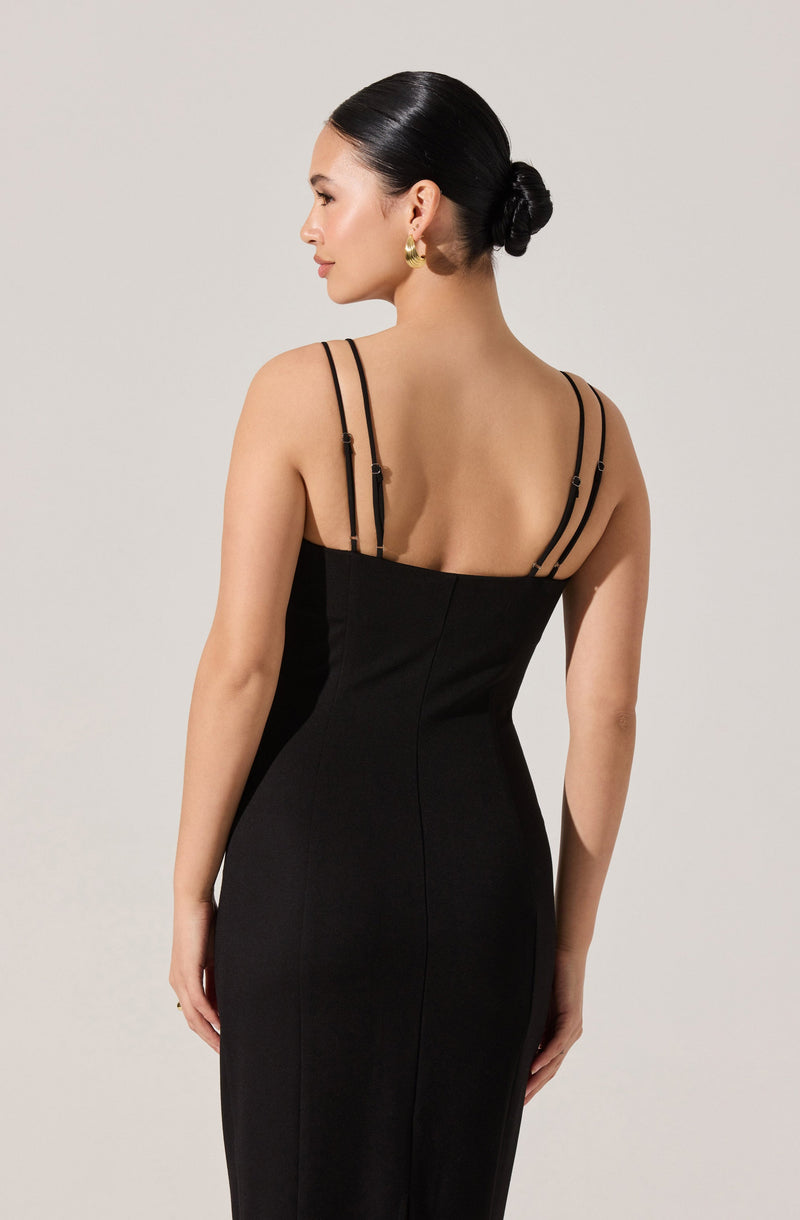 A woman is wearing a fitted black dress with spaghetti straps that have adjustable buckles. The dress is sleeveless and has a sleek, elegant appearance. The woman has her hair styled in a low bun and is wearing large gold hoop earrings.