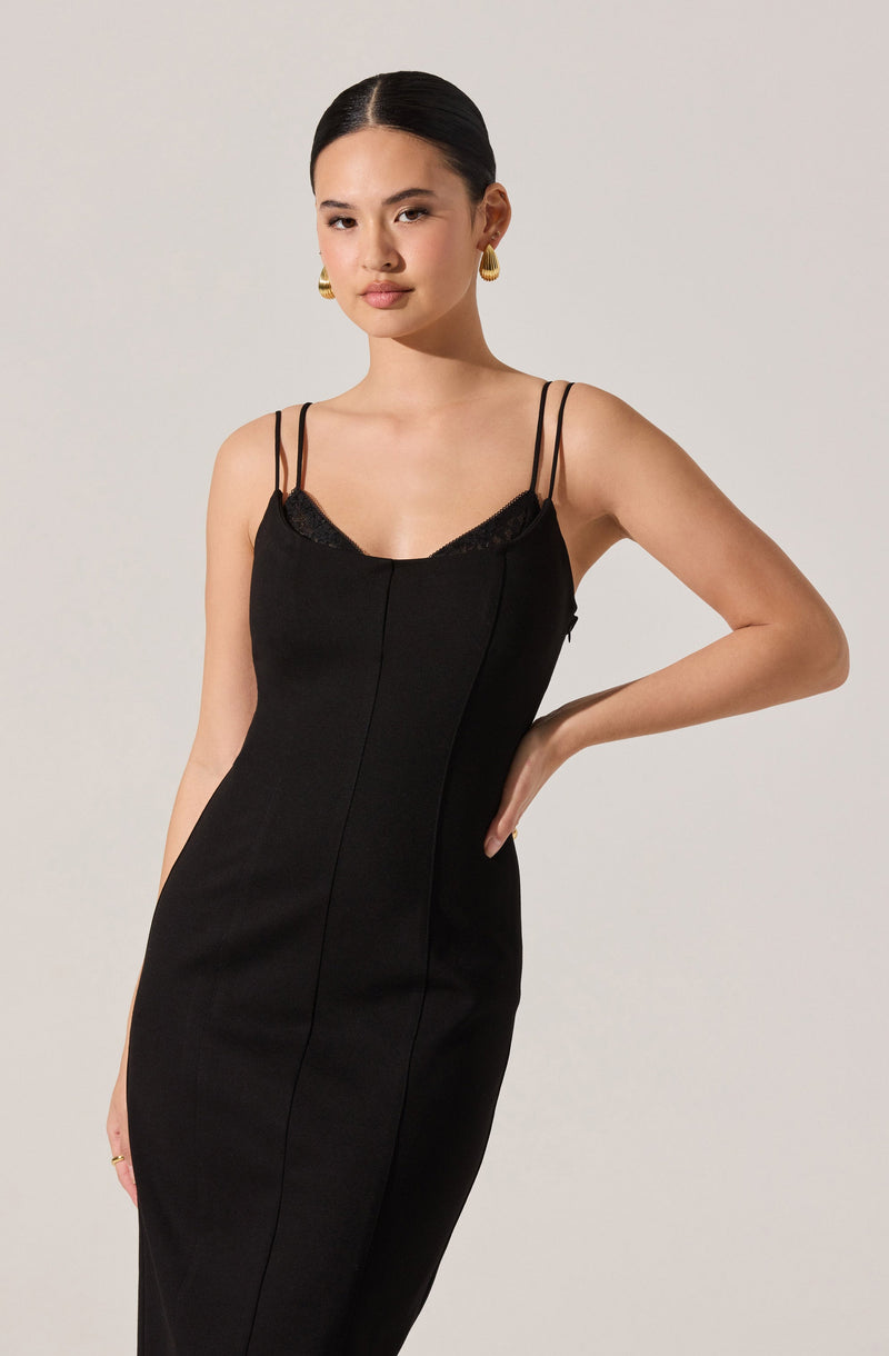A woman wearing a black, fitted dress with thin double shoulder straps. The dress has a slight V-neckline with lace trim detail. She is accessorized with large, gold hoop earrings.