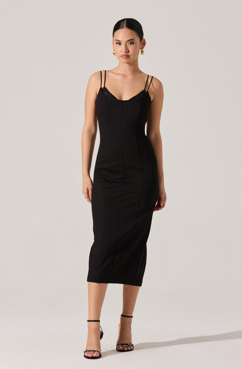 A woman is wearing a fitted, ankle-length black dress with thin spaghetti straps. The dress features subtle lace detailing at the top and has vertical seam lines running down the front. She is also wearing black high-heeled sandals with ankle straps and gold earrings.
