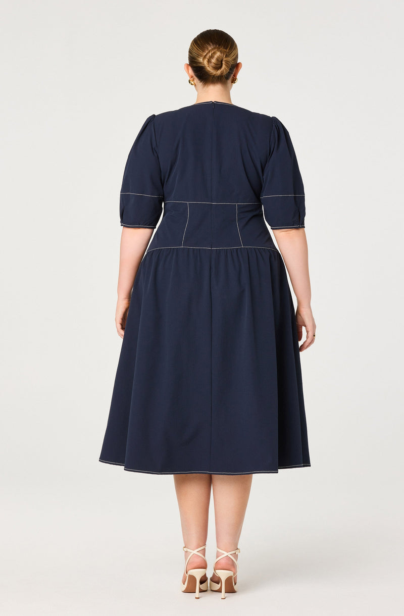 A woman wearing a navy blue dress with puffed short sleeves, a fitted bodice with visible white topstitching, and a gathered midi-length skirt. She is also wearing beige high-heeled shoes with ankle straps.