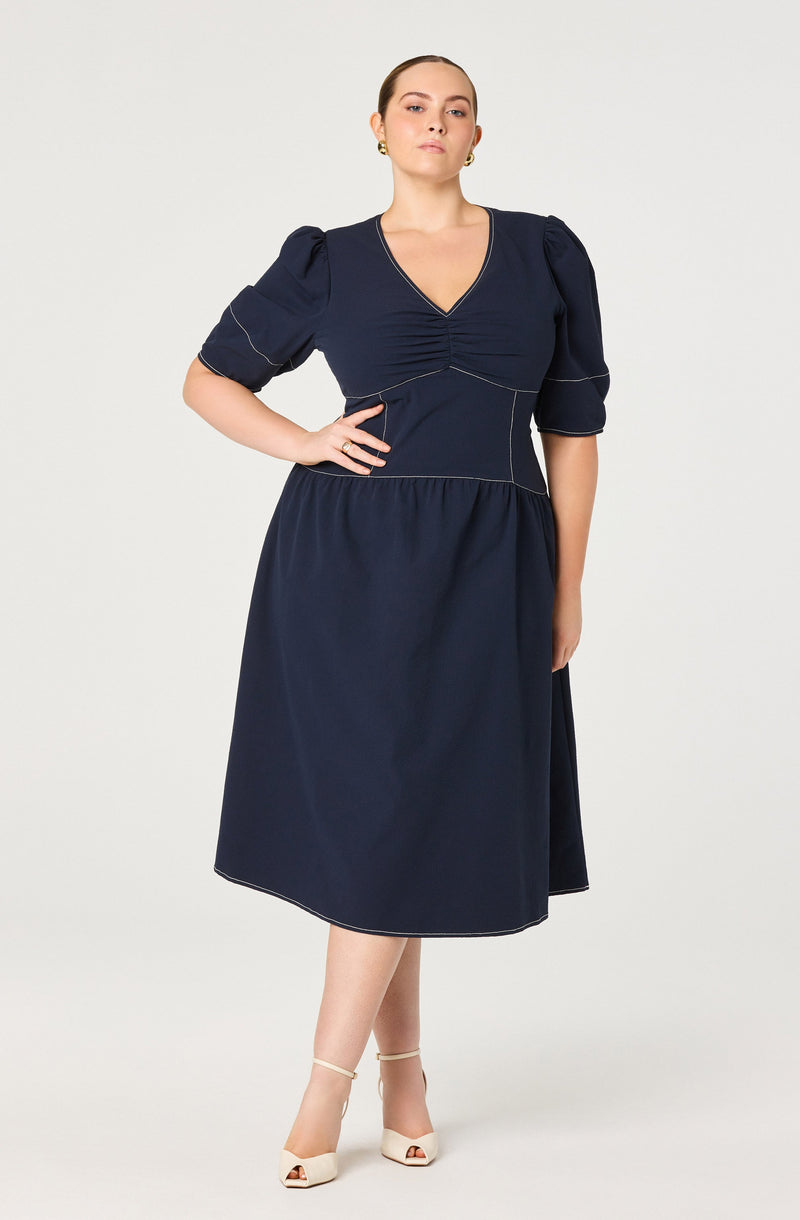A woman is wearing a navy blue dress with a fitted bodice and a V-neckline. The dress features puffed, elbow-length sleeves with subtle piping details and a waistband that creates a gathered effect at the skirt. The skirt is A-line and midi-length, falling below the knees. She is also wearing light-colored open-toe heels with ankle straps.