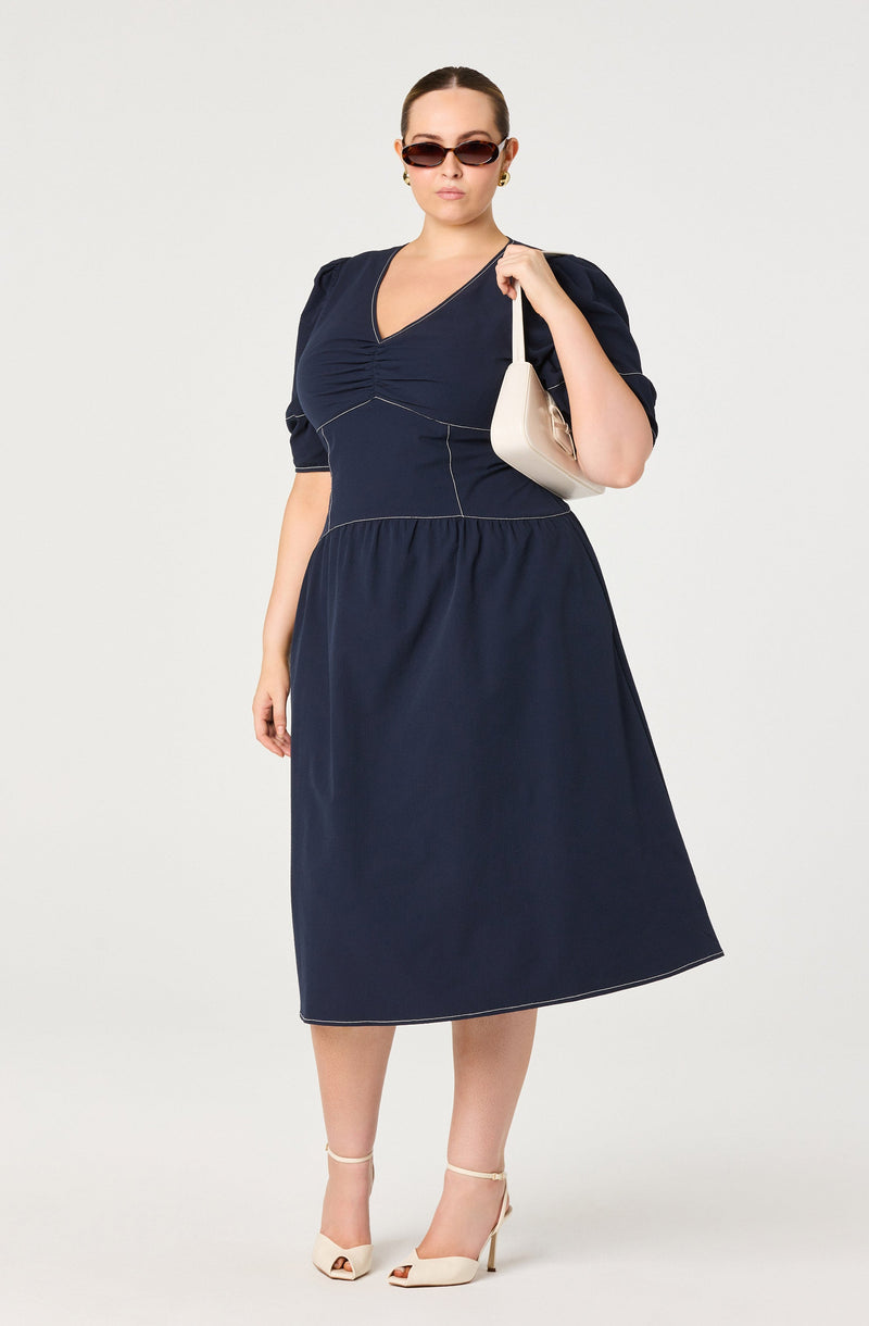 A woman is wearing a navy blue midi dress with puff sleeves and a V-neckline. She has on dark sunglasses and earrings, and she is carrying a white handbag over her shoulder. She is also wearing white high-heeled sandals.