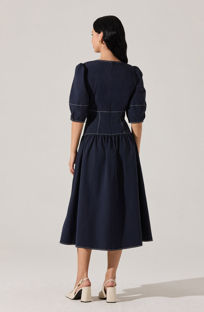 A woman is wearing a navy blue dress with puffed three-quarter-length sleeves, a fitted waist with white topstitching details, and a gathered, full-length skirt. She pairs the dress with light-colored, high-heeled sandals that have ankle straps.