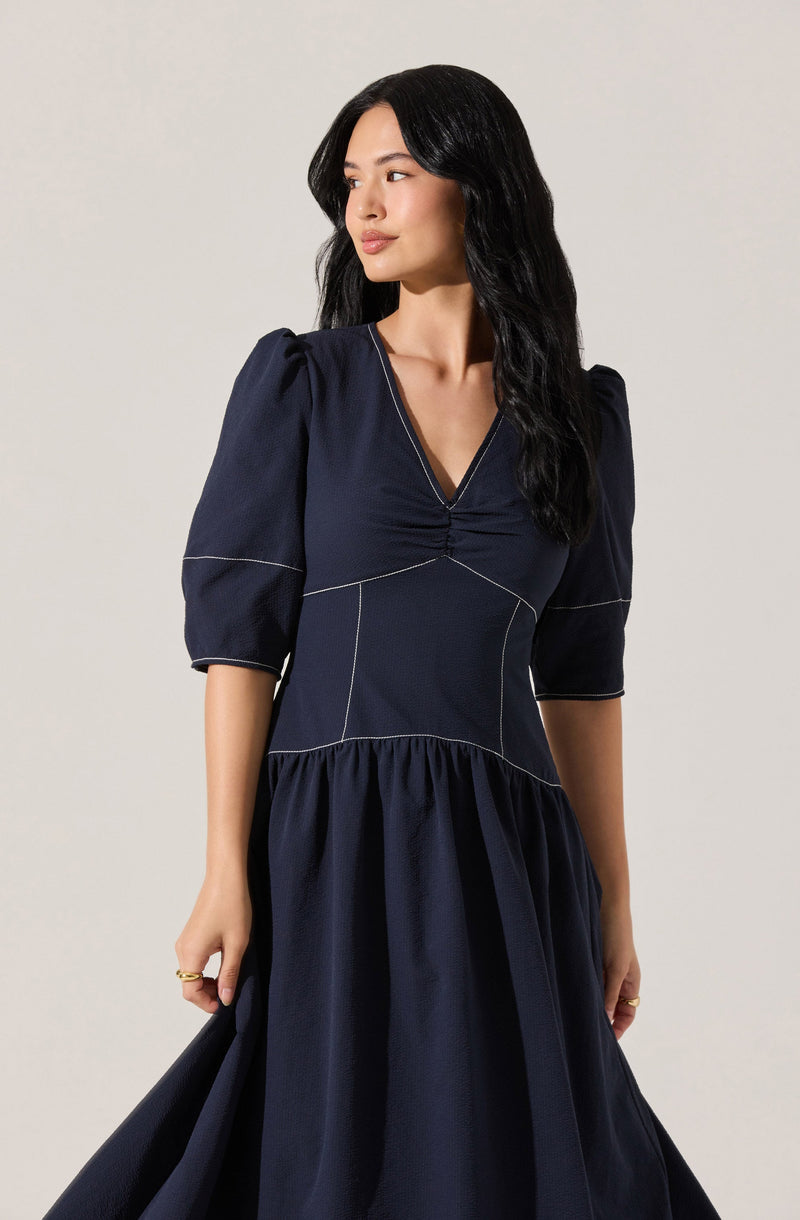 A woman is wearing a navy blue dress with a fitted bodice and a gathered, full skirt. The dress features puffed, three-quarter length sleeves with subtle stripe detail along the seams and a V-neckline with a ruched center. The fabric appears smooth and slightly shiny.