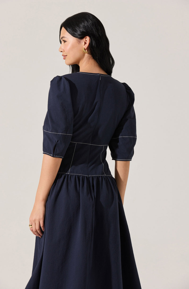 A woman is wearing a navy blue dress with puffed, three-quarter length sleeves that have visible white stitching details. The dress has a fitted waistline with a gathered, full skirt. The fabric appears to be a slightly textured material.