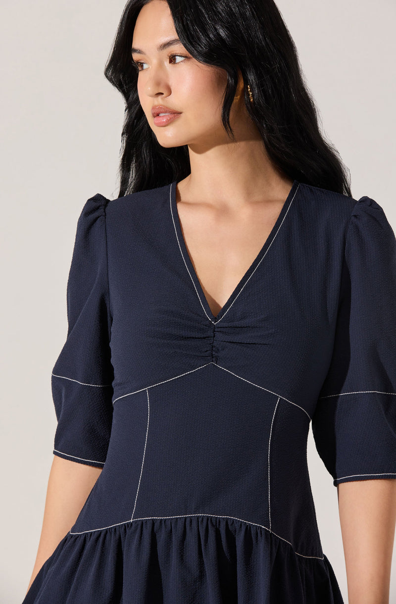 A woman is wearing a dark navy blue dress with a V-neckline and puffed sleeves. The dress features visible white stitching that creates geometric panels and a gathered detail at the center of the bust. The skirt of the dress has a ruffled, gathered hemline.
