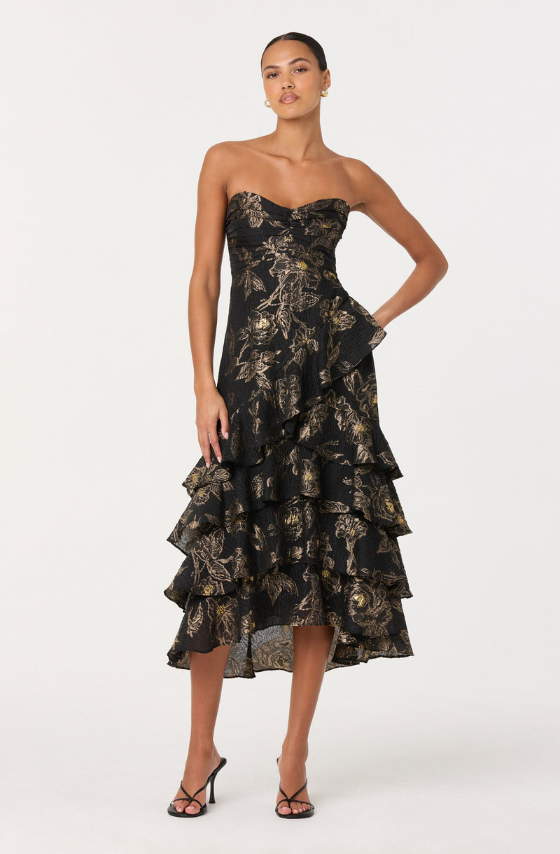 A woman is wearing a strapless, black dress with a gold floral pattern. The dress features a fitted bodice and multiple layered ruffles cascading down the skirt, which reaches mid-calf length. She completes the look with black high-heeled sandals.