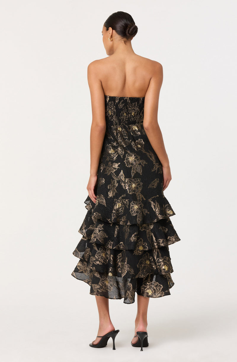 A woman is wearing a strapless dress with a fitted bodice and a tiered, ruffled skirt. The dress features a black background with a gold and beige floral pattern. She is also wearing black high-heeled sandals.