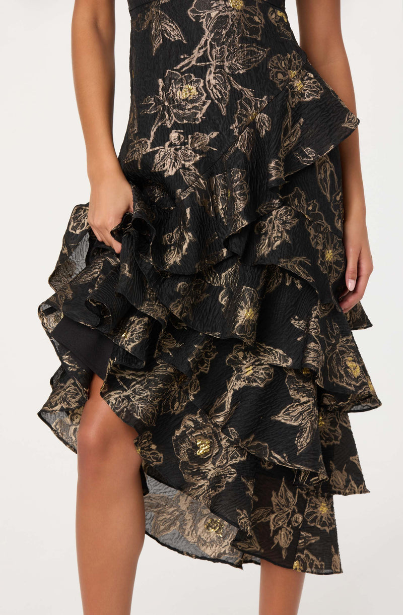 A close-up view of a black dress with a textured, floral pattern in beige and gold. The dress features multiple layered ruffles with a slightly ruffled hem, creating a voluminous and elegant appearance. The fabric has a crinkled texture, adding depth to the floral design. The person wearing the dress is holding one of the ruffled layers with their left hand, and their right hand is resting at their side. The dress appears to be knee-length or slightly above the knee.